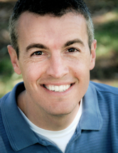 A photograph of a smiling man with dark eyebrows and greying hair wearing a blue polo shirt on top of a white T-shirt looking at the viewer