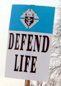 A photograph of a placard at the March of Life that reads "Defend life" on the bottom with the Emblem of the Order in a blue band on top.