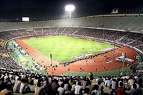 An open-roof sport arena at the nighttime, with grass field (at the centre) and athletic track (around the field), sitting pavilions full with crowd watching a football match