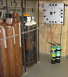 A panel on the wall is connected to diving cylinders by hoses. Nearby are several much larger cylinders, some painted brown and others black