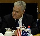 Man with grey hair and a dark suit, white shirt, dark tie sitting down, looking to his right. In front of him, a coffee pot, a bottle of water, and a small U.S. flag.