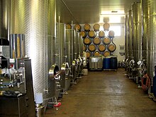 Colour photo showing stainless steel fermentation tanks in the UK. The tanks are located on both sides of an aisle at the bottom of which oak barrels for aging, stacked on a bracket against the wall can be seen. The floor is painted and pitched to a drainage channel in the middle of the winery.