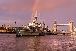 The bow of a large blue warship, moored on a river, with a bridge in the background.