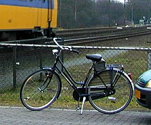 Farbfoto. Dunkles Hollandrad, tiefer Einstieg, Beleuchtung, Klingel, Gepäckträger, keine Handbremse, keine Gangschaltung. Im Hintergrund Gleise.