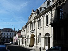 Photo d'une rue en enfilade, avec sur la droite la façade ensoleillée du théâtre le Palace, présentant un fronton sculpté et un grand porche.