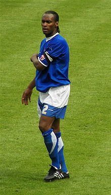 A black footballer in blue-and-white shorts, shirt and socks standing on a football pitch with his left arm across his chest