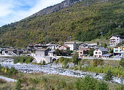 Photographie en couleurs d'un village au bas d'une montagne, le long d'un cours d'eau quasiment à sec.