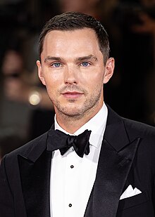 A young, Caucasian man with short, dark hair and facial stubble wearing a black tuxedo jacket paired with a white pocket square, a white shirt with black buttons and a black bow tie, as he looks directly into the camera.