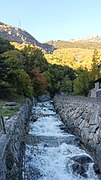 Photographie d'un torrent agité, l'eau étant plus ou moins gelée, contenue dans le canal du torrent en pierres, avec du soleil derrière ainsi que de la forêt.