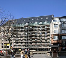 Eingerüstete Fassade eines siebengeschossigen Hotelgebäudes von der Domplatte aus fotografiert, blauer Himmel, im Vordergrund eine kleine Menschengruppe