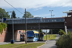 en T-banestasjon lagt i bro over en vei. Veien er delt inn i veibane, trikketrasé og fotgjengerfelt, fra venstre til høyre. Både en sporvogn og et T-banesett er synlig på bildet.