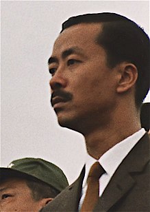 A middle-aged man with side-parted black hair and a moustache, in a black suit, white shirt and brown tie. To the left is a clean-shaven Asian man with black hair and a green military cap.