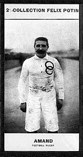 Portrait en demi grandeur d'un homme moustachu en maillot blanc avec les anneaux olympiques brodés sur la poitrine.