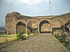 Outdoor picture of an atrium with a cobbled path leading to a stone structure with five arches. An altar is carved into the wall behind the central arch.