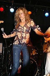 A woman with long blonde hair wearing a floral-patterned shirt and jeans holding a microphone on a stage