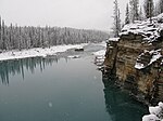 A river flows by a cliff and through a snow-covered forest.