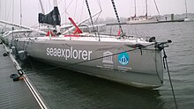 Vu de l'avant, bateau gris endommagé, amarré au ponton. On remarque notamment le bout-dehors cassé, le balcon avant arraché et des éraflures sur le côté tribord de la coque.