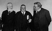 Three men in black suits against a plain wall; the leftmost man is slender and has a white beard, the middle man is carrying a large tome under his arm and an umbrella in hand, and the rightmost man is obese with small glasses and a moustache