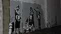 A gray wall is painted with black and white images. A 1980s style boom box sits on the ground. Older adults are dressed in 1980s street style clothing.