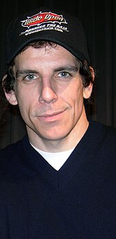 Stiller is facing the camera and smiling. He is wearing a baseball cap and a blue shirt with a white t-shirt underneath.