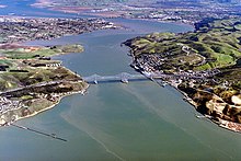 Rolling hills covered partially with buildings and roads frame the banks of a large channel of opaque water, which connects two larger bodies of water at the top left and bottom.