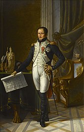 Painting shows a slightly balding man with dark hair standing in a room holding a building plan. He wears an early 1800s French military uniform: a blue coat with a white waist coat, white breeches, and black boots which rise above his knees.