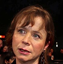 Emily Watson steht auf einem roten Teppich. Im Hintergrund ist ein Absperrzaun zu sehen, an dem einige Personen stehen. Watson trägt ein schwarzes Oberteil, an beiden Ohren je einen kleinen und einen großen, durch einen Haken miteinander verbundenen schwarzen Ohrring sowie ihre roten Haare als bis zum Hals reichende Bobfrisur mit Pony.