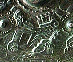 Detail of a metal object showing embossed scene of a horse-drawn chariot.