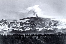 Black and white photograph of Mount Wrangell showing a plume rising above the mountain peak and dark material coating the slopes.