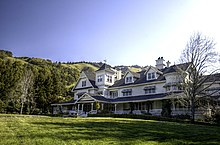 Une grande maison blanche derrière laquelle on aperçoit des collines couvertes de forêts