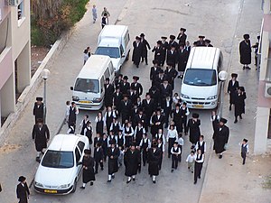 A group of people clad mostly in black, some partially in white, walking along a street between several white vehicles, seen from above and in front