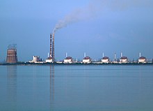 Die Gebäuden sind mittig der Farbfotografie aneinandergereiht und liegen an einem Fluss, der bei klarem Himmel widerspiegelt. Der Kühlturm und beide Kamine befinden sich auf der linken Seite, auf der rechten Seite sind sechs identische kleine Gebäude.