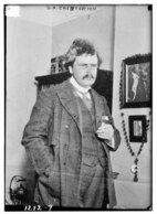 A plump man with small glasses, a moustache, and unkempt hair smokes a cigarette in a tweed suit indoors