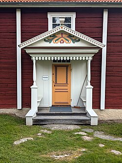 Vitmålad förstukvist med sadeltak och en utsmyckning på krönet. Förstukvisten ramar in en gulmålad dörr och pryds av dekorationer i rött, gult och grönt.