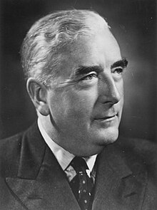 A middle-aged, portly man with wavy greying hair sits half profile with a dark suit and tie, with a ceremonial handkerchief in his suit breastpocket.