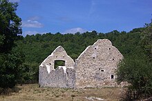 Photo montrant les restes d'une grande grange en pierre calcaire blanche : quatre murs et deux pignons. Le ciel est bleu, l'herbe jaunie est grillée par le soleil.