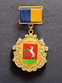 Medal of honor granted to the city of Lublin that has the form of a cartouche with a brick crown on a background of acanthus leaves. The cartouche contains the coat of arms of the city.