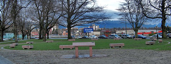 In a park, 14 coffin-like benches of pink stone are set in a circle. A higher slanted pink panel is visible in the foreground