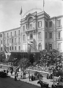 Period photo of a hotel, with four stories, large square windows, and a wrought iron portico with flags. Pedestrians, horse-drawn carriages and a motor car are before it