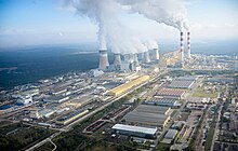 Aerial view of the Belchatow Power Station site, with smoke coming from its smokestacks, and surrounding buildings.