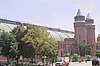 A large round-roofed building running the width of the image behind a city street and trees. At the right are two conical towers and an entrance pavilion.