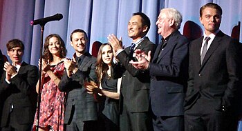 A man in a black suit, a woman in a pink dress, a man in a plaid suit, a woman in a black dress, a Japanese man in a black suit, and an old man in a blue suit clap their hands, while a man in a black suit stands. A microphone stand is in the foreground, and blue curtains are in the background.