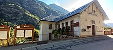 Photographie de la mairie d'Orelle en été avec des fleurs au centre dudit chef-lieu, il y a des panneaux municipaux avec l'inscription d'entrée révélant que le bâtiment est la mairie.