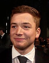 Taron Egerton steht auf einem roten Teppich. Im Hintergrund ist ein Absperrzaun zu sehen, an dem einige Personen stehen. Egerton trägt ein schwarzes Jackett, ein weißes Hemd, eine schwarze Krawatte sowie seine braunen Haare bis zu den Ohren rasiert, oben flach und nach rechts gescheitelt.