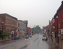 A wide, wet street with tall commercial buildings on either side. Two approaching vehicles have their headlights on, and a traffic light behind them is green. In the rear of the image the street narrows to go over a small bridge.