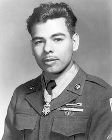 Head and shoulders of a bald man with dark beard, with a medal hanging from a blue ribbon around his neck.