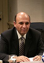 Man with a bald head, some dark hair at the back, sitting down, looking straight ahead, wearing a dark brown jacket, beige shirt, brown and beige striped tie. His hands are on the table in front of him, as is a glass of water.