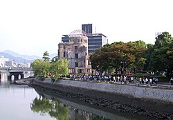 A shot along a river. There is a bridge in the distance, and a ruined domed building in the middle distance. People walk along the footpath that runs parallel to the river