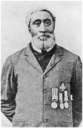 A black-and-white photograph of a man with a beard and short hair looking at the viewer and wearing a button-up jacket displaying four medals