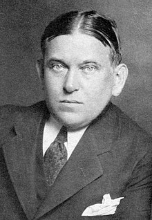 A black and white photographic portrait of critic H. L. Mencken. His hair is parted in the middle, and he appears to be leaning on his left arm. He is wearing a dark tie and a dark suit with peak lapels. A white handkerchief is visible in his suit pocket.